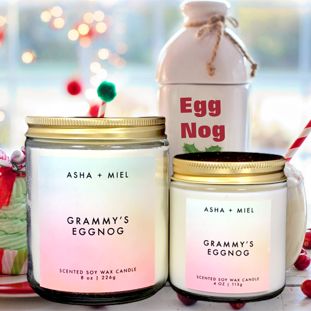 One 8 ounce and one 4 ounce Grammy's Eggnog Candle in glass jars with gold tops on background of table with Egg Nog, berried and a cupcake